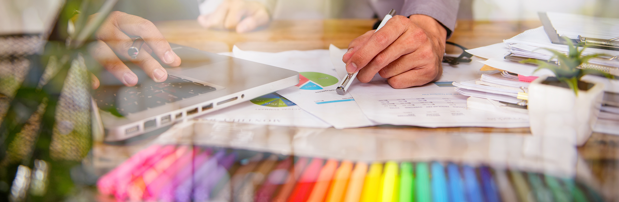 A man's hand pointing to a document with a stylus, surrounded by a stack of papers and colored pencils.