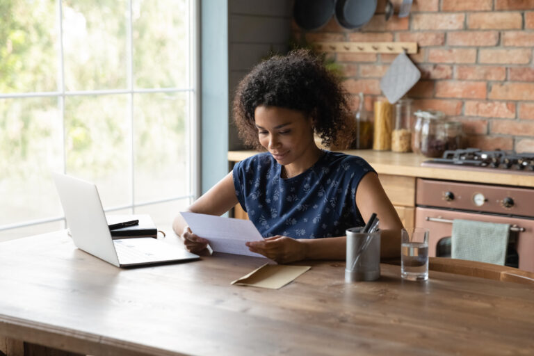 A woman reading a letter, sitting at a table with a laptop at home